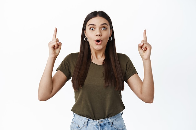 Impresionada chica morena jadeando asombrada, señalando con el dedo hacia arriba y mirando emocionada al frente, mirando la oferta promocional, de pie sobre una pared blanca