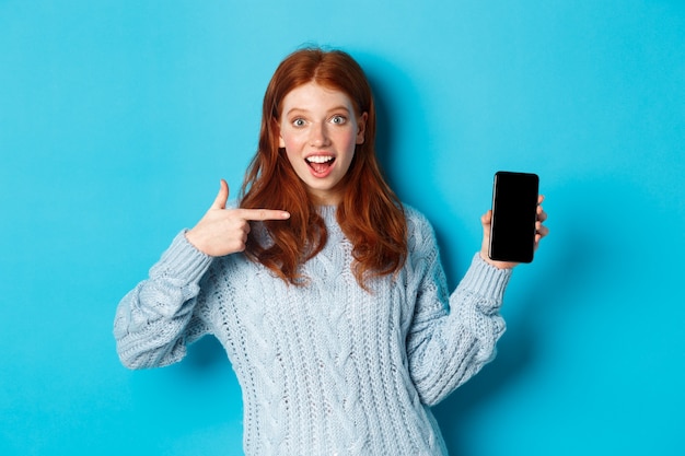 Impresionada chica pelirroja apuntando a la pantalla del teléfono, mostrando la aplicación de teléfono inteligente u oferta en línea y sonriendo emocionada, de pie en suéter sobre fondo azul.