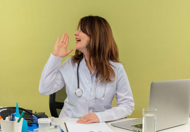 Impresionada doctora de mediana edad con bata médica y estetoscopio sentado en el escritorio con herramientas médicas y computadora portátil girando de cabeza a lado llamando a alguien aislado