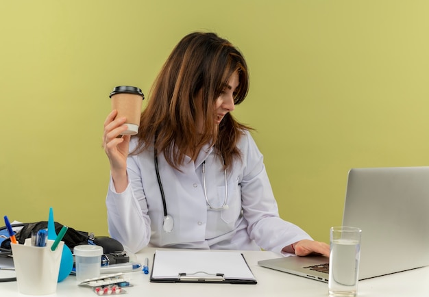 Impresionada doctora de mediana edad con bata médica y estetoscopio sentado en el escritorio con herramientas médicas y portapapeles usando una computadora portátil sosteniendo una taza de café de plástico aislada