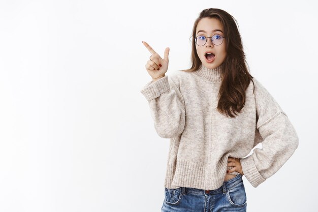 Impresionada, emocionada y sorprendida, atractiva mujer de los años 20 con gafas y un suéter cálido que se deja boquiabierto de asombro.