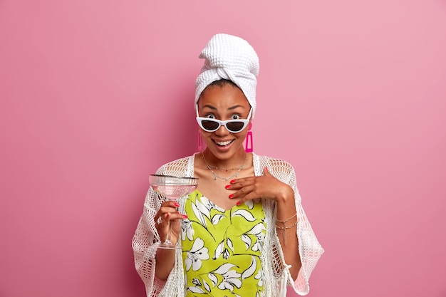 Impresionada feliz modelo de mujer étnica de piel oscura sostiene un cóctel alcohólico, usa elegantes gafas de sol, una toalla de baño envuelta en la cabeza, pasa tiempo libre en una fiesta de pijamas, posa contra una pared rosada