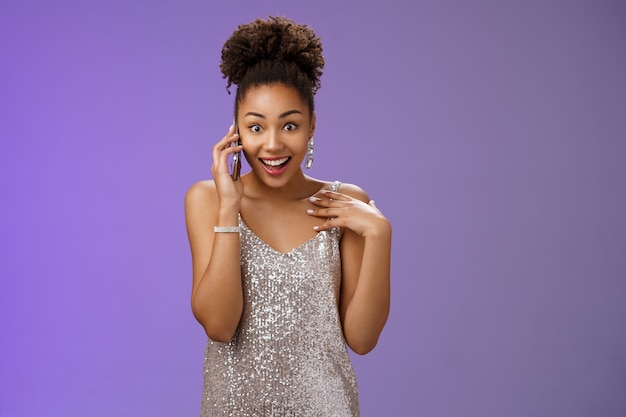 Impresionada habladora encantadora mujer afroamericana en vestido de noche plateado brillante apuntando a sí misma divertido sorprendido sonriendo hablando smartphone abrir ojos asombrados, de pie fondo azul.