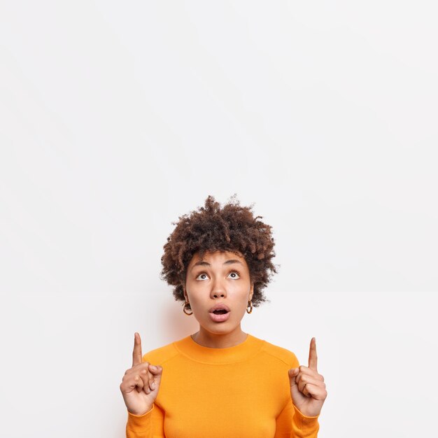 Impresionada hermosa mujer afroamericana se ve sorprendida arriba indica en el espacio de la copia muestra una oferta increíble sorprendida por las grandes ventas viste ropa casual aislada sobre una pared blanca. Promoción