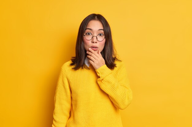 Impresionada hermosa mujer asiática sostiene la barbilla se ve atónita oye algo impactante usa suéter casual de gafas redondas.