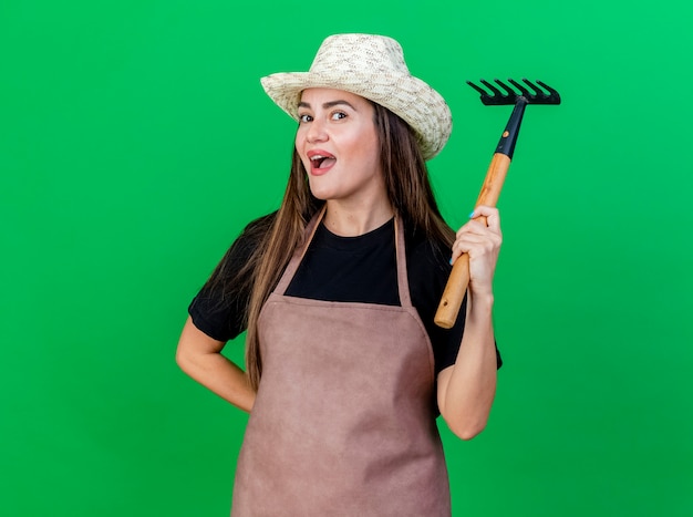 Foto gratuita impresionada hermosa niña jardinero en uniforme con sombrero de jardinería levantando rastrillo aislado en verde