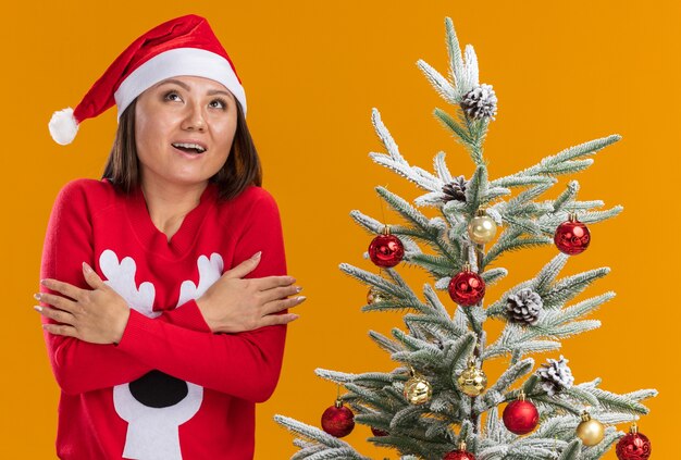 Impresionada joven asiática vistiendo gorro de navidad con suéter de pie cerca del árbol de navidad cruzando las manos aisladas sobre fondo naranja