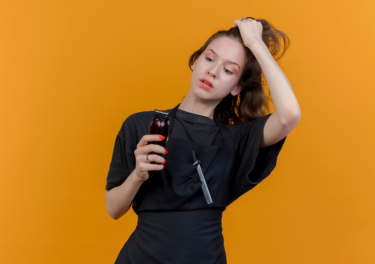 Foto gratuito impresionada joven barbero eslavo vistiendo uniforme sosteniendo y mirando cortapelos y tirando del cabello aislado sobre fondo naranja con espacio de copia