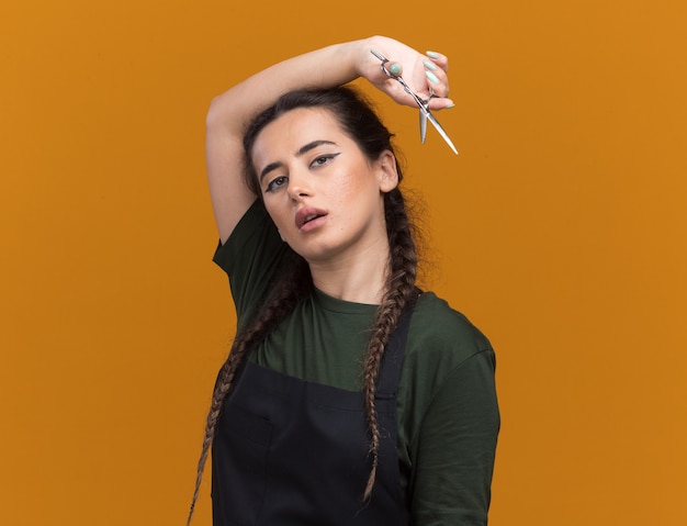 Foto gratuita impresionada joven barbero en uniforme sosteniendo unas tijeras alrededor del hombro aislado en la pared naranja con espacio de copia