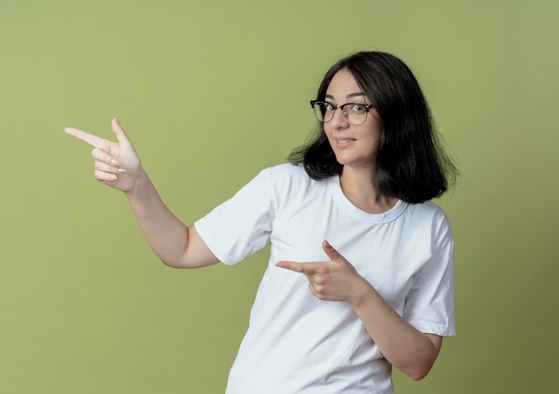 Impresionada joven bastante caucásica con gafas apuntando al lado aislado sobre fondo verde oliva