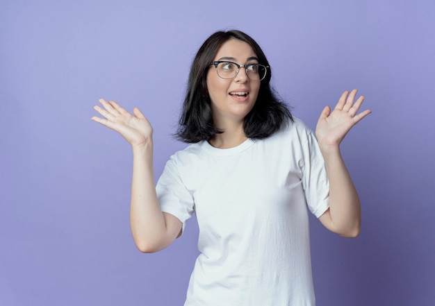 Impresionada joven bastante caucásica con gafas mirando a un lado y mostrando las manos vacías aisladas sobre fondo púrpura con espacio de copia