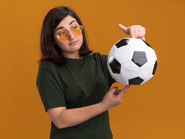 Impresionada joven bastante caucásica en gafas de sol sosteniendo y mirando la bola aislada en la pared naranja con espacio de copia