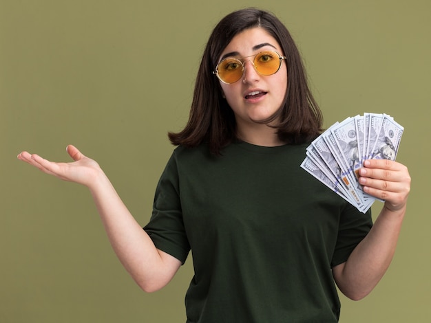 Impresionada joven bastante caucásica en gafas de sol tiene dinero y mantiene la mano abierta en verde oliva