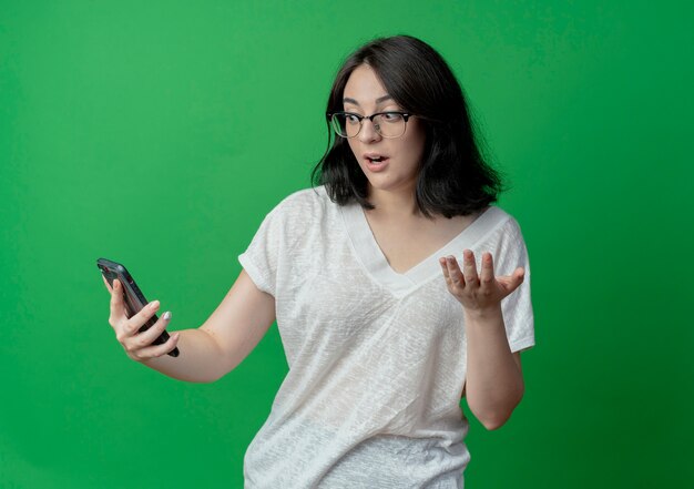Impresionada joven bastante caucásica con gafas sosteniendo y mirando el teléfono móvil y mostrando la mano vacía aislada sobre fondo verde