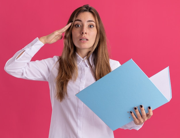 Impresionada joven bastante caucásica haciendo gesto de saludo y sostiene la carpeta de archivos en rosa