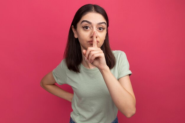 Impresionada joven bastante caucásica manteniendo la mano detrás de la espalda haciendo gesto de silencio aislado en la pared rosa con espacio de copia