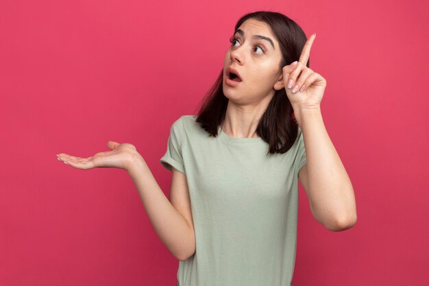 Impresionada joven bastante caucásica mostrando la mano vacía mirando al lado apuntando hacia arriba aislado en la pared rosa