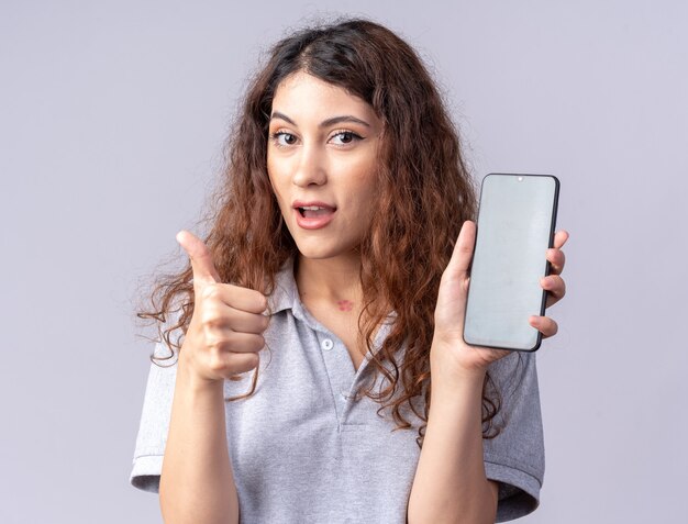 Impresionada joven bastante caucásica mostrando teléfono móvil y mostrando el pulgar hacia arriba