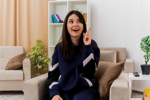 Impresionada joven bastante caucásica sentada en un sillón en la sala de estar diseñada mirando de lado y levantando el dedo