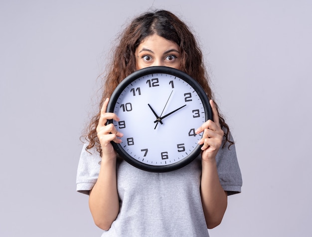 Impresionada joven bastante caucásica sosteniendo el reloj detrás de él aislado en la pared blanca con espacio de copia