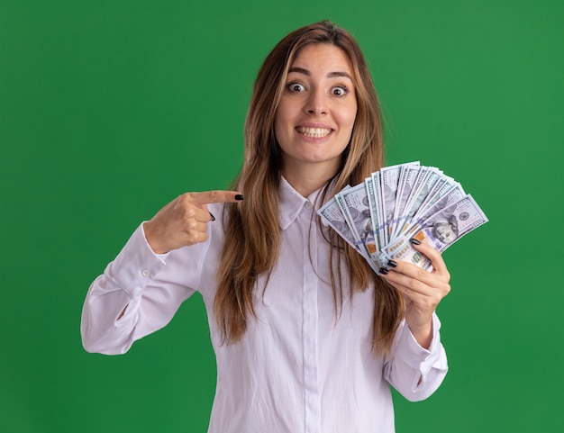 Foto gratuita impresionada joven bastante caucásica sostiene y apunta al dinero aislado en la pared verde con espacio de copia