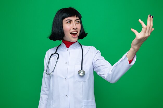 Impresionada joven bastante caucásica en uniforme médico con estetoscopio apuntando y mirando al lado