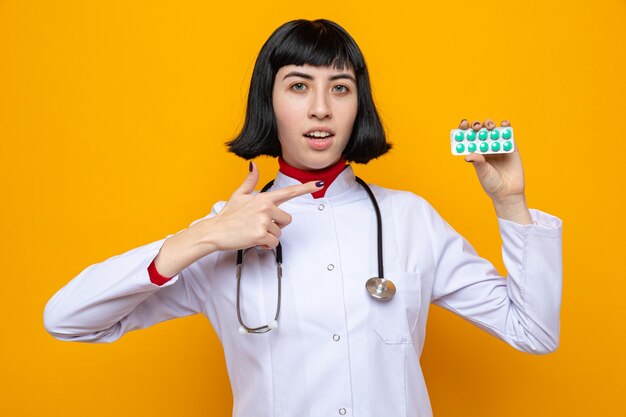 Impresionada joven bastante caucásica en uniforme médico con estetoscopio sosteniendo y apuntando al envase de la píldora