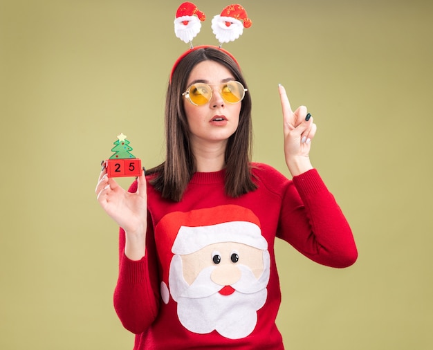 Foto gratuita impresionada joven bastante caucásica vistiendo suéter de santa claus y diadema con gafas sosteniendo el juguete del árbol de navidad con fecha mirando al lado apuntando hacia arriba aislado sobre fondo verde oliva