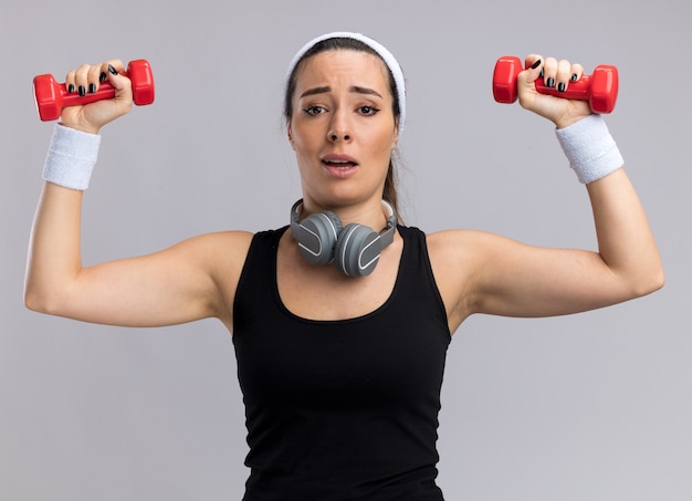 Impresionada joven bastante deportiva con diadema y muñequeras levantando pesas con auriculares alrededor del cuello aislado en la pared blanca