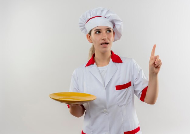 Impresionada joven bonita cocinera en uniforme de chef sosteniendo plato vacío mirando y apuntando hacia arriba aislado en la pared blanca