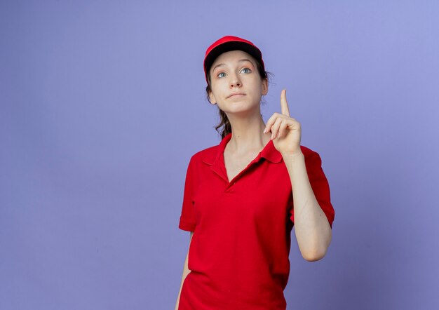 Impresionada joven bonita entrega mujer vistiendo uniforme rojo y gorra levantando el dedo mirando hacia arriba
