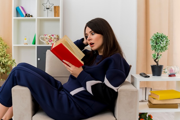 Impresionada joven bonita mujer caucásica sentada en un sillón en la sala de estar diseñada libro de lectura poniendo la mano en la cabeza