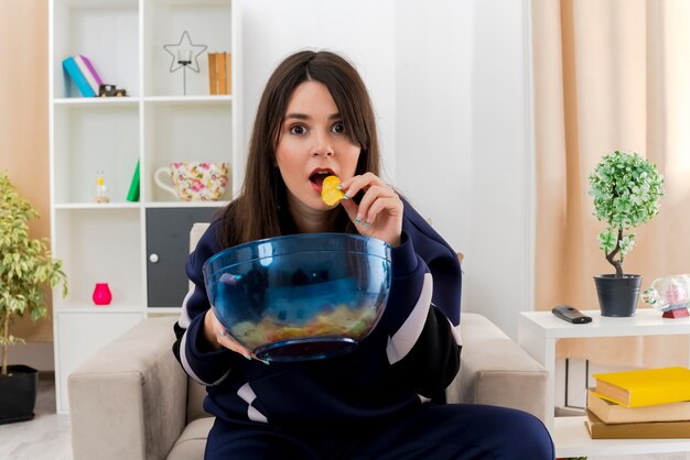 Impresionada joven bonita mujer caucásica sentada en un sillón en la sala de estar diseñada sosteniendo un tazón de papas fritas mirando y comiendo papas fritas