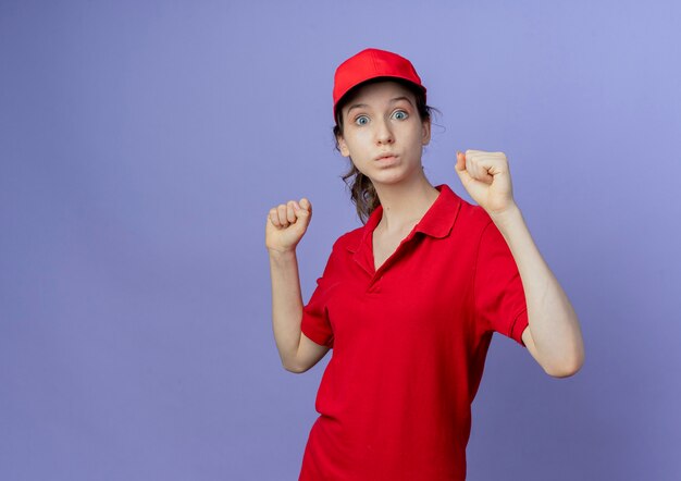 Impresionada joven bonita mujer de entrega con uniforme rojo y gorra levantando los puños