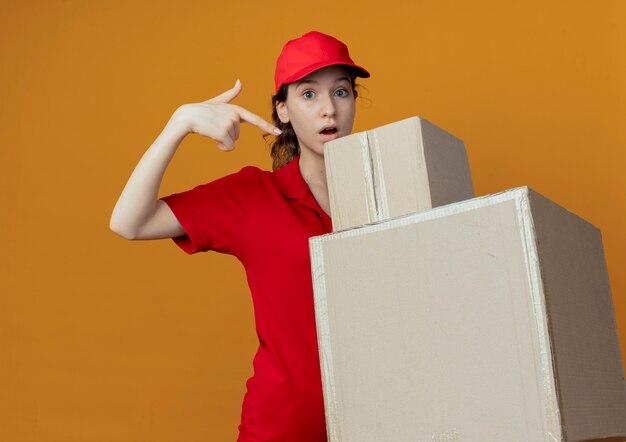 Impresionada joven bonita mujer de entrega con uniforme rojo y gorra sosteniendo y apuntando a cajas de cartón