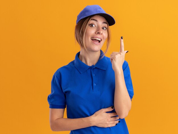 Impresionada joven bonita repartidora en uniforme apunta hacia arriba en naranja