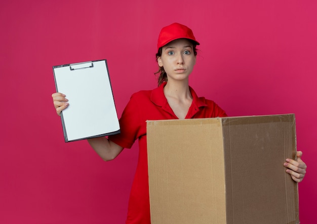 Impresionada joven bonita repartidora en uniforme rojo y gorra con caja de cartón y portapapeles aislado sobre fondo carmesí