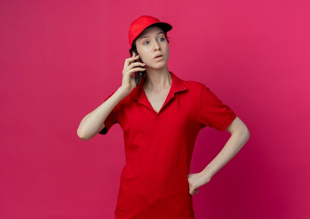Impresionada joven bonita repartidora en uniforme rojo y gorra hablando por teléfono mirando al lado con la mano en la cintura aislada sobre fondo carmesí con espacio de copia