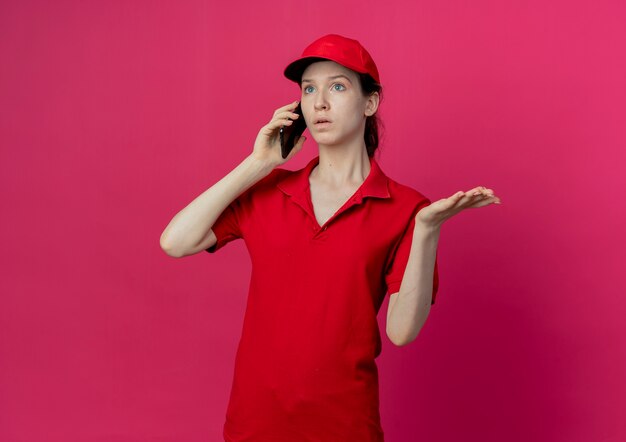 Impresionada joven bonita repartidora en uniforme rojo y gorra hablando por teléfono mirando directamente y mostrando la mano vacía aislada sobre fondo carmesí con espacio de copia