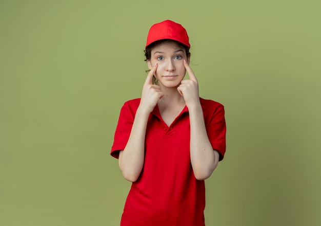 Impresionada joven bonita repartidora en uniforme rojo y gorra poniendo los dedos debajo de los ojos aislados sobre fondo verde oliva con espacio de copia