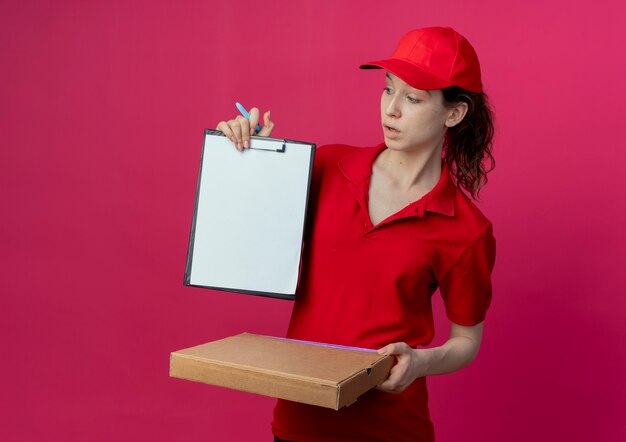 Impresionada joven bonita repartidora en uniforme rojo y gorra sosteniendo el bolígrafo del paquete de pizza y mostrando el portapapeles mirando el portapapeles aislado sobre fondo carmesí con espacio de copia