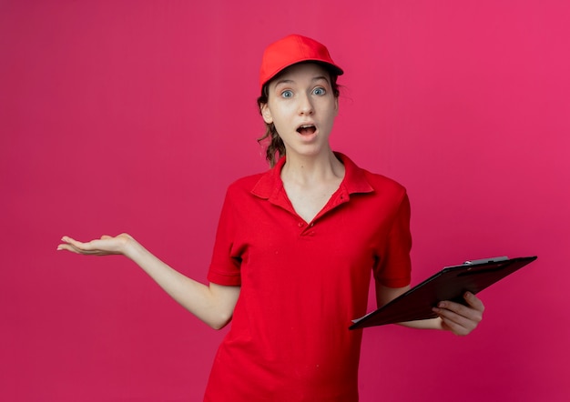 Impresionada joven bonita repartidora en uniforme rojo y gorra sosteniendo el portapapeles y mostrando la mano vacía aislada sobre fondo carmesí