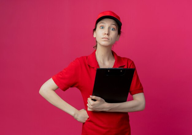 Impresionada joven bonita repartidora en uniforme rojo y gorra sosteniendo el portapapeles y poniendo la mano en la cintura aislada sobre fondo carmesí con espacio de copia