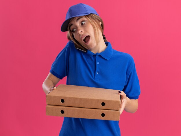 Impresionada joven bonita repartidora en uniforme sostiene cajas de pizza y habla por teléfono en rosa