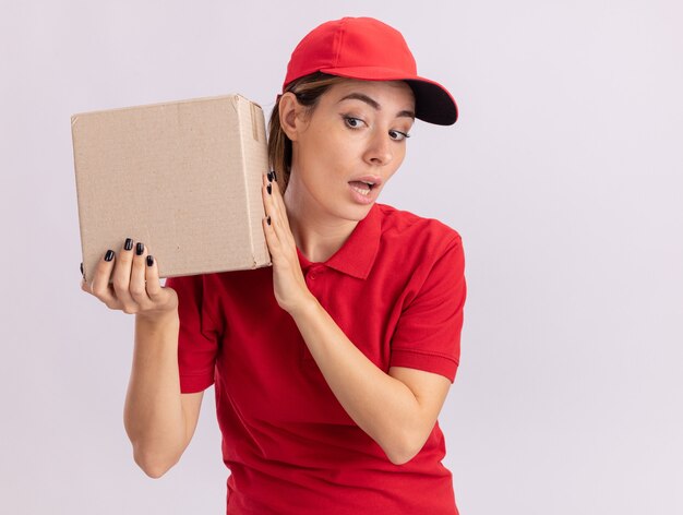 Impresionada joven bonita repartidora en uniforme sostiene cardbox cerca de la oreja aislada en la pared blanca