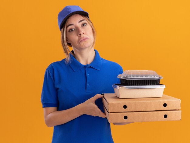 Impresionada joven bonita repartidora en uniforme sostiene paquetes de alimentos de papel y contenedores en cajas de pizza en naranja