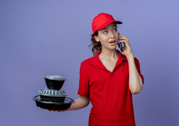 Impresionada joven bonita repartidora vestida con uniforme rojo y gorra mirando al lado sosteniendo contenedores de comida y hablando por teléfono aislado sobre fondo púrpura