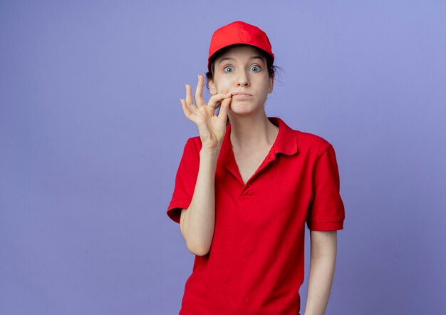 Impresionada joven bonita repartidora vestida con uniforme rojo y gorra mirando a la cámara cerrando la boca aislada sobre fondo púrpura con espacio de copia