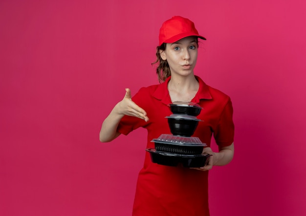 Impresionada joven bonita repartidora vestida con uniforme rojo y gorra sosteniendo y apuntando con la mano a los recipientes de comida aislados sobre fondo carmesí con espacio de copia
