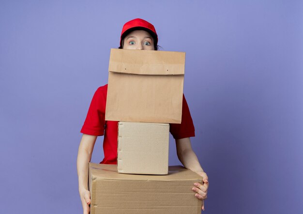 Impresionada joven bonita repartidora vestida con uniforme rojo y gorra sosteniendo cajas de cartón y paquete de papel y mirando desde detrás del paquete de papel